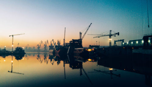 Silhouette cranes at dock reflection in sea against clear sky