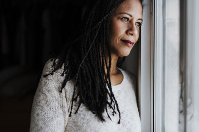 Contemplating woman looking through window while standing at home