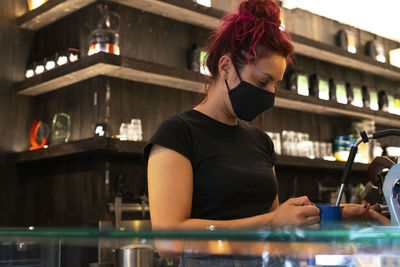Side view of female bartender in face mask standing at counter in coffee shop and making delicious hot beverage in coffee machine during coronavirus epidemic