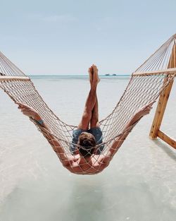 Rear view of man relaxing on hammock at beach against sky