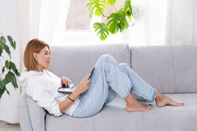 Young or middle age woman sitting with laptop on grey couch in home office with monstera plant.