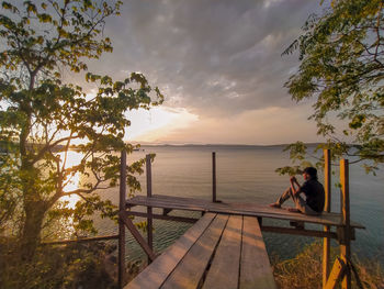 People sitting on railing by sea against sky