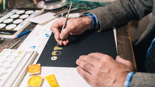 Side view crop anonymous male artist creating jewelry ring design and coloring sketch with yellow paint while working in professional workshop