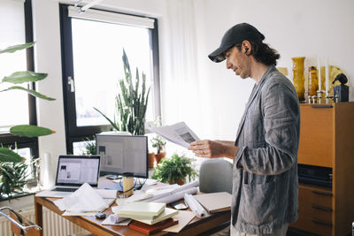 Side view of male architect analyzing blueprint while standing by desk in home office