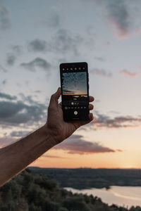 Hand holding smart phone against sky during sunset