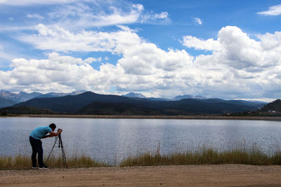 Side view of man photographing on lakeshore against cloudy sky