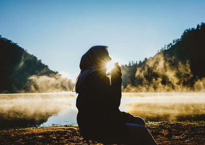 Woman sitting by lake against sky during sunset