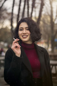 Portrait of smiling young woman holding cigarette standing against trees