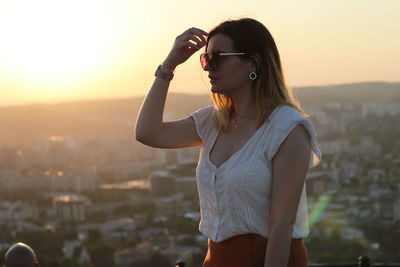 Young woman wearing sunglasses standing against cityscape during sunset