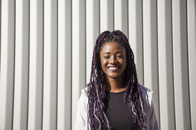 Happy young african american female with long dyed braids smiling brightly and looking at camera while having fun against gray wall in sunlight