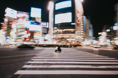 Person riding motor scooter on road in illuminated city at night