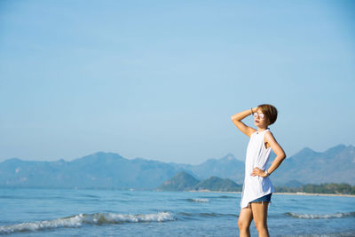 Young woman with hand on hip standing at beach against clear blue sky