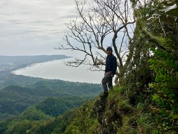 Man standing on rock by bare tree on mountain against sky