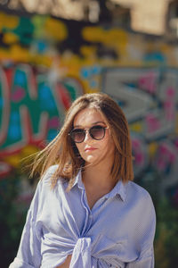 Portrait of beautiful young woman wearing sunglasses while standing against graffiti wall