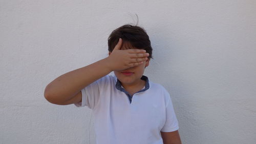 Boy covering eyes with hand against white wall