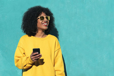 Smiling young woman wearing sunglasses using mobile phone against wall