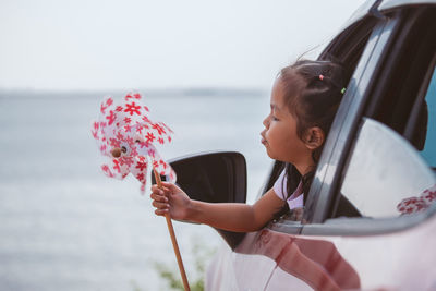 Cute girl holding pinwheel toy while leaning on car window at beach