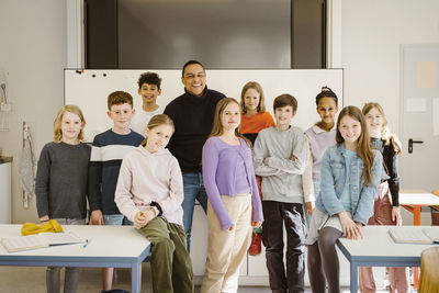 Portrait of smiling multiracial students standing with male teacher by desk in classroom