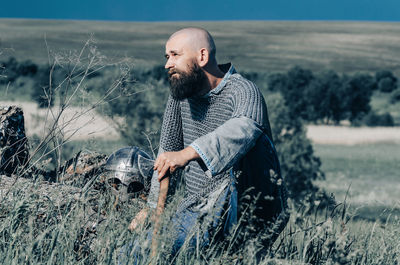 Viking. bald bearded man in metal chain mail over linen shirt sits on hill, leaning on ax. 