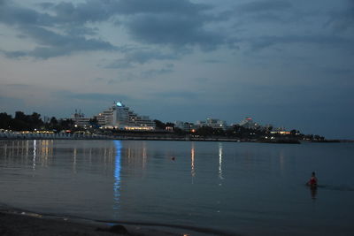 Illuminated buildings by sea against sky at dusk