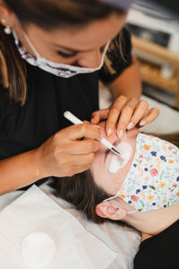 High angle of crop anonymous female master in medical mask applying foaming cleanser on eyelashes of client after extension in modern salon