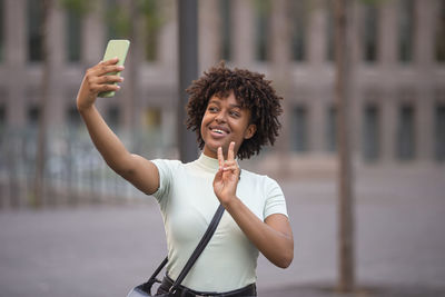 Beautiful young woman with curly hair and fashionable clothes takes a selfie outdoors in the city.