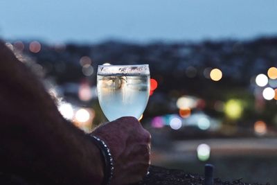 Close-up of man hand holding drink against illuminated city at night