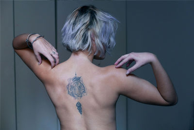 Rear view of topless young woman with tattoo standing against wall