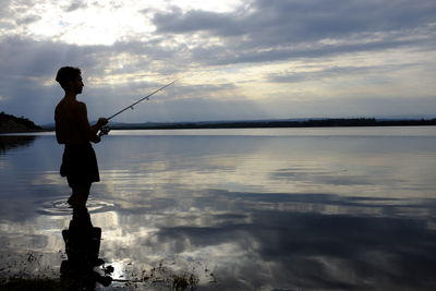 Silhouette man fishing in lake against cloudy sky during sunset