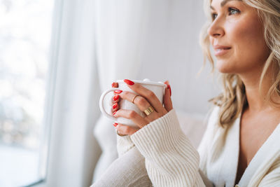 Young woman in white cardigan with cup of cocoa in hands looking at window with winter landscape 