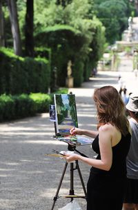 Rear view of woman painting while standing on footpath at park