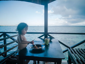 Girl sitting on table by sea against sky