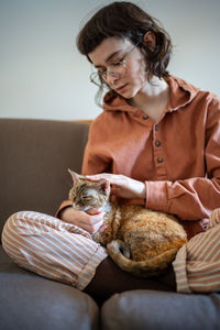 Caring pet owner young woman stroking petting cat sitting on couch at home enjoying communication.