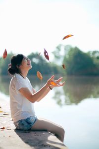 Side view of woman catching autumn leaves while sitting by lake