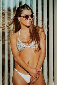 Young woman in bikini wearing sunglasses standing against fence