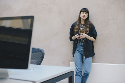 Young woman using phone while standing against wall