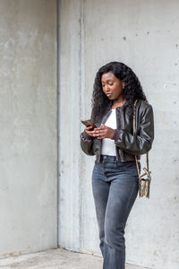 Young woman using mobile phone while standing against wall
