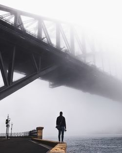 Rear view of man standing on wall under bridge over sea against sky