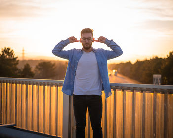 Full length portrait of man standing on railing against sky during sunset