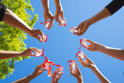 Cropped hands of friends toasting drinks against clear blue sky