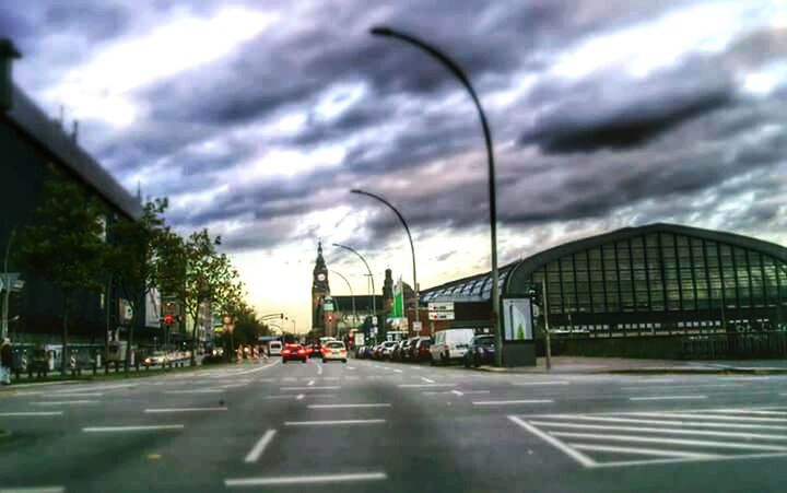 sky, transportation, architecture, cloud - sky, building exterior, built structure, road, the way forward, car, cloudy, street, city, land vehicle, road marking, cloud, diminishing perspective, mode of transport, street light, incidental people, vanishing point