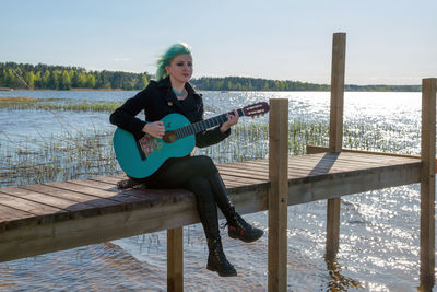 Hipster young woman with turquoise guitar sitting on pier over lake