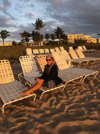 Portrait of senior woman wearing sunglasses sitting on deck chair at beach against cloudy sky during sunset