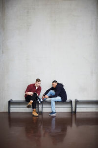 Full length of male friends with technologies sitting on bench against wall in university