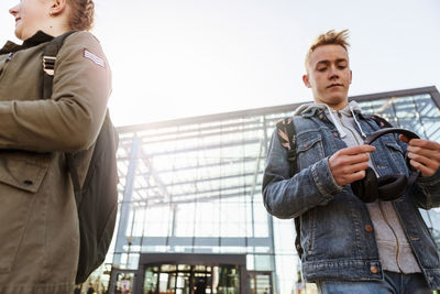 Low angle view of teenage boy holding headphones while standing with friend in city