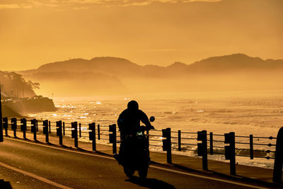 Silhouette person on motorcycle by railing against sky and sea during sunrise