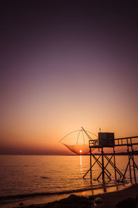 Silhouette fishing net over sea against orange clear sky during sunset