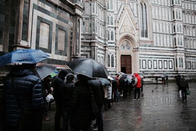 People with umbrellas by historic building on street in city during rainy season