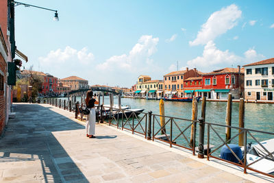 Woman photographing canal amidst buildings in city against sky