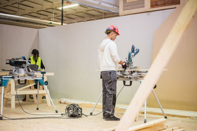 Full length of teenage carpenter cutting plank with electric saw while standing at workshop